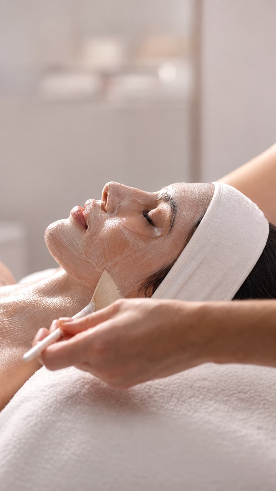Applicazione con il pennello di una maschera peeling sul viso di una donna sdraiata con una fascia per capelli